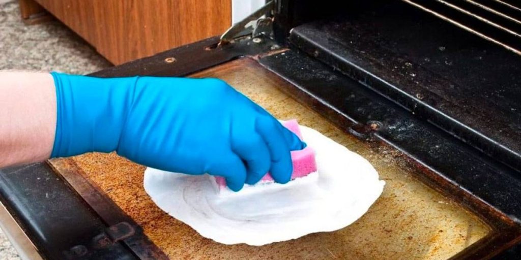 A hand wearing a blue disposable glove using a sponge to scrub the inside of a glass oven door.