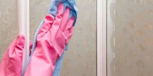 Hands wearing pink rubber gloves cleaning a glass shower door with a blue microfiber cloth.